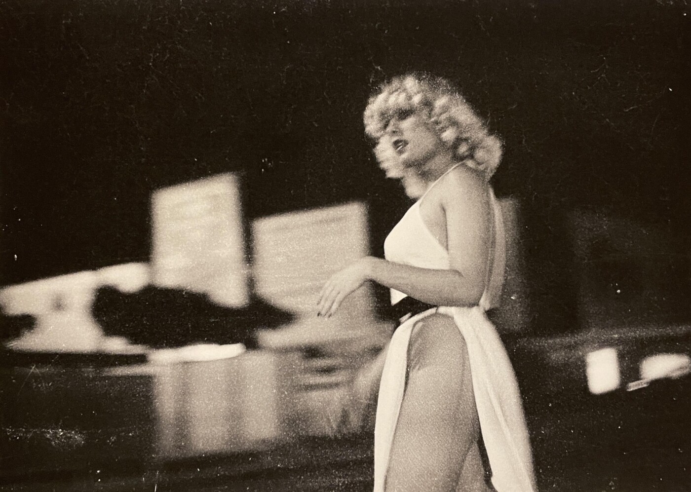 Black-and-white photograph of trans woman standing in the street in a low-cut white dress with a slit in the leg.