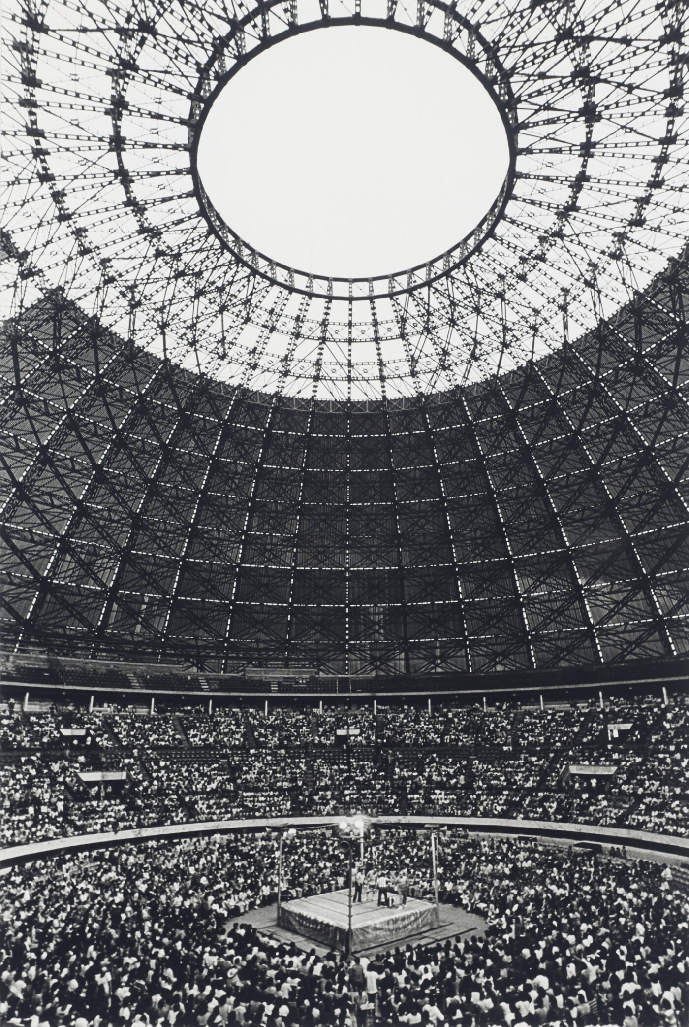 A black-and-white vertical photograph depicts a domed open-air arena shot from within. Two thirds of the composition are dominated by the concentric metal armatures of the arena’s architecture. At the center of the top edge of the photograph is an oculus through which light pours into the space. The audience fills the bottom third of the photograph, their attention fixed on a wrestling ring centered on the bottom edge.
