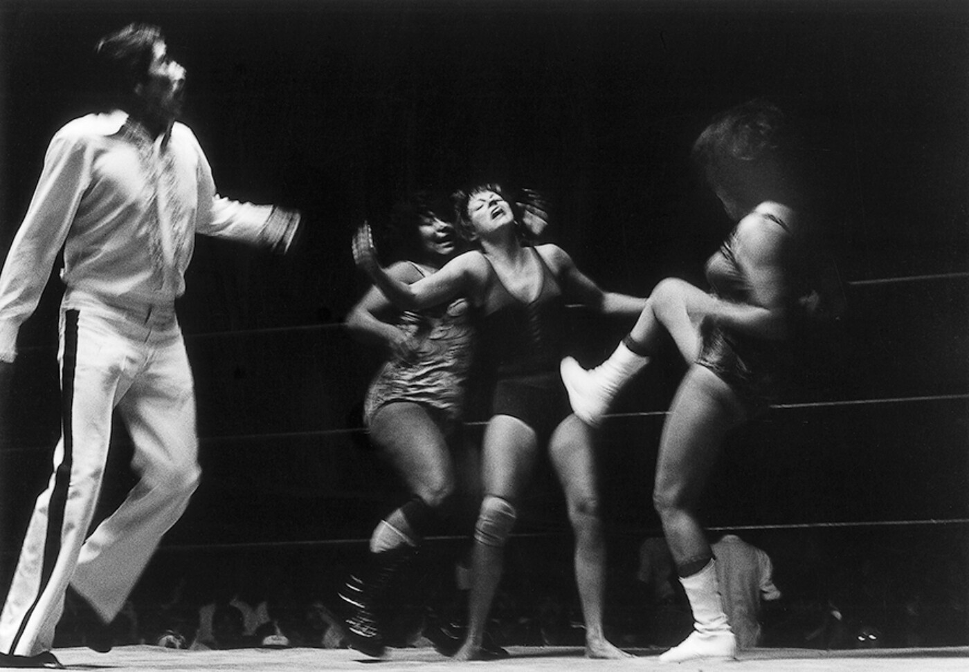 A black-and-white horizontal photograph depicts four figures against a saturated black background. At center, a woman wearing a dark leotard stands with legs spread and arms thrown out, slightly blurry with motion. Her head is cast back, and her arms are open. Behind her, a second woman seems to run up to support her, one hand reaching for her head, another coming up to support her arm. Right of them, a third woman leans on her left leg, while her right white-booted foot kicks out at the stomach of the first woman. At the left edge, a man dressed in white springs into the frame, his bent legs suggesting that he is mid-jump. The drooping horizontal lines of two ropes can be seen on the right edge of the photograph, suggesting that the action takes place in a wrestling ring.