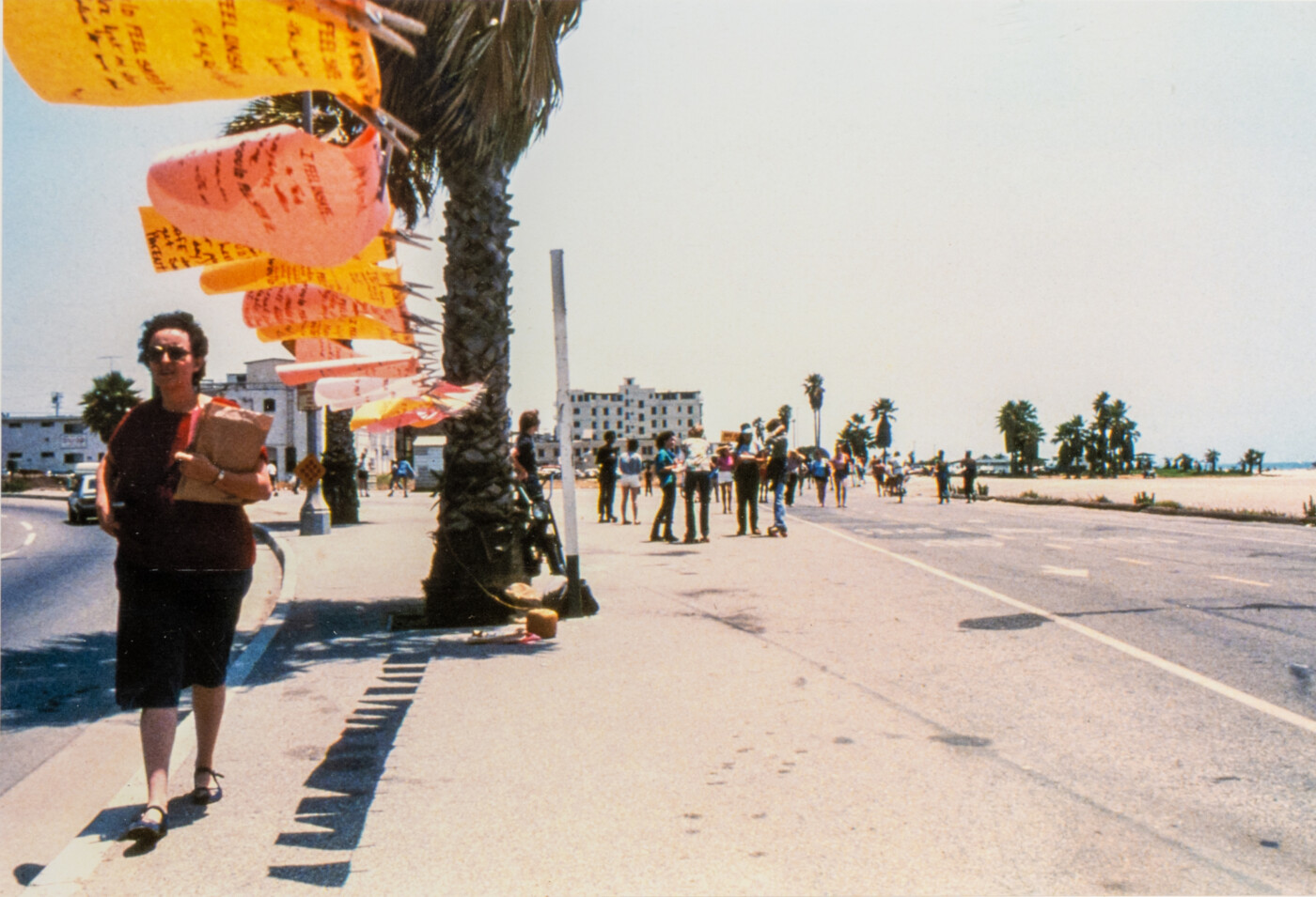 A color photograph in which a woman holding a brown paper bag walks down a sidewalk, towards the camera. Above her, pink and orange rectangles of cloth inscribed with text are suspended from a clothesline. The clothesline is tethered on one end to a palm tree visible in the foreground of the image, left of center; the other disappears in the upper left edge of the photo. To the woman’s right is a street with cars, and to her left is a paved beach-side walkway with pedestrians and runners.
