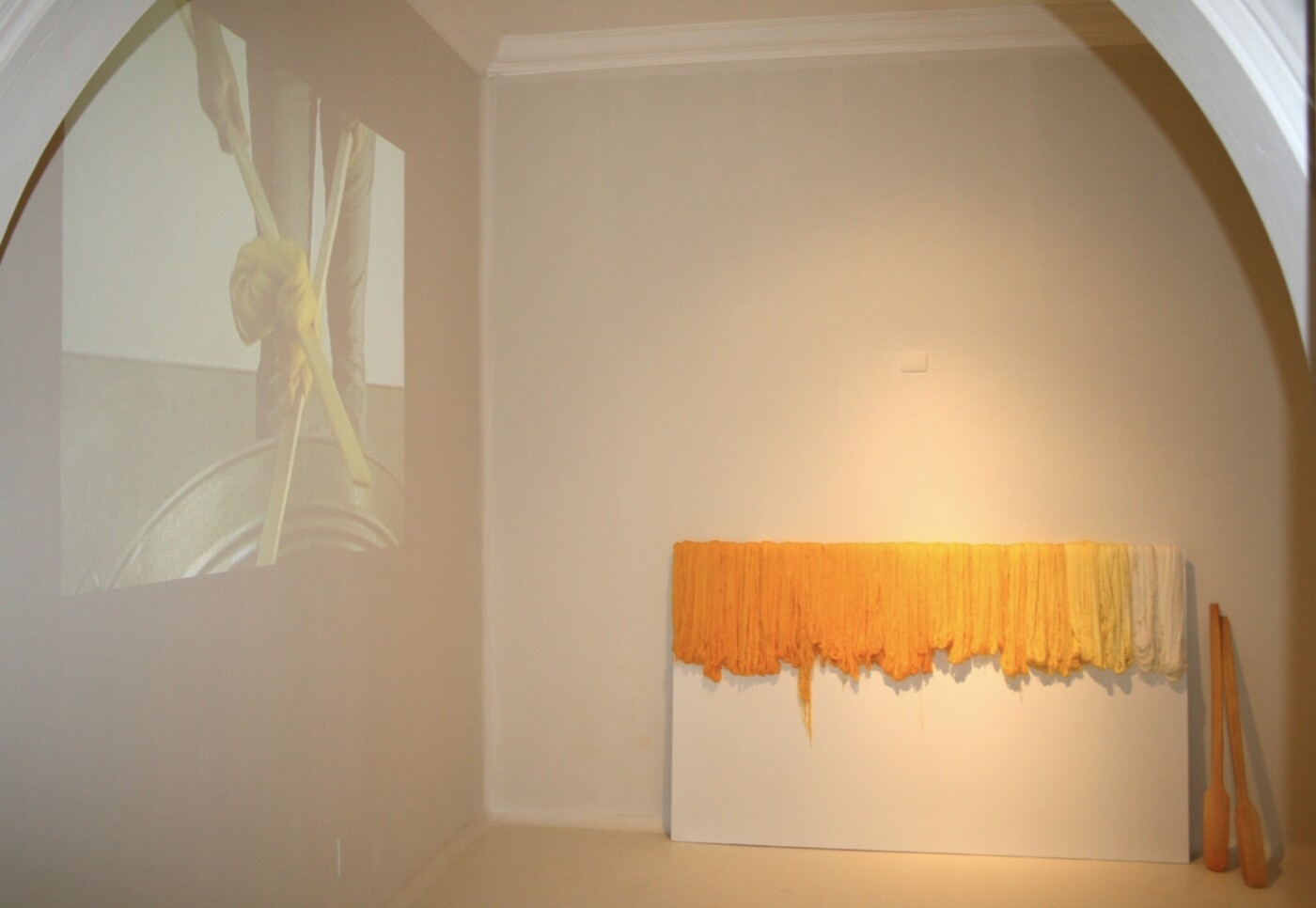 Installation artwork containing a leaning panel at center, draped with a fabric fringe in shades of yellow, a set of paddle-shaped wooden objects on the righthand side of the panel, and a video projection on a lefthand wall.