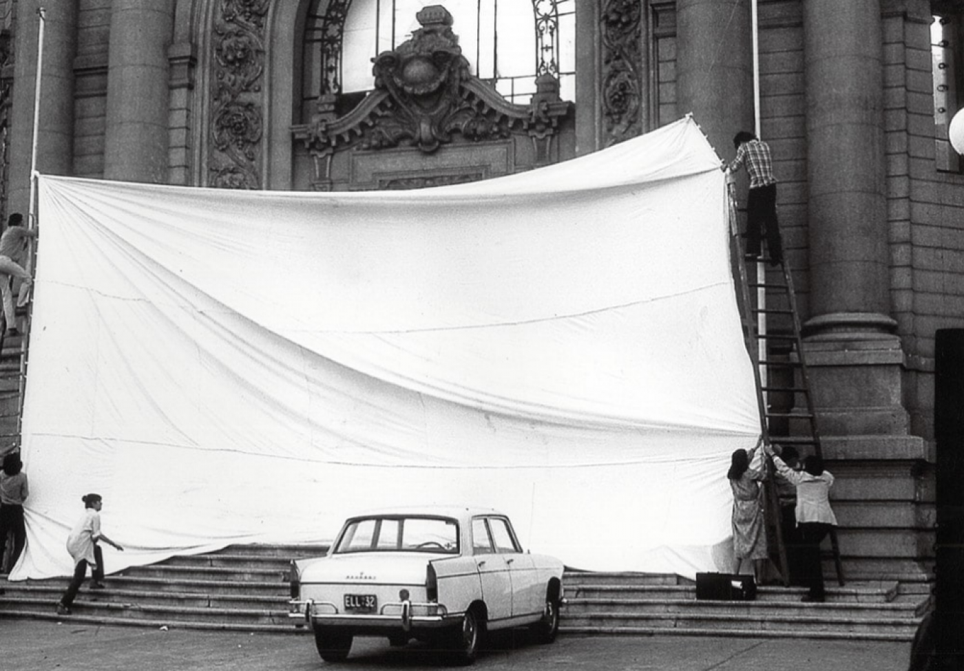 Black and white photograph of a people installing a large white sheet over a large building entrance.