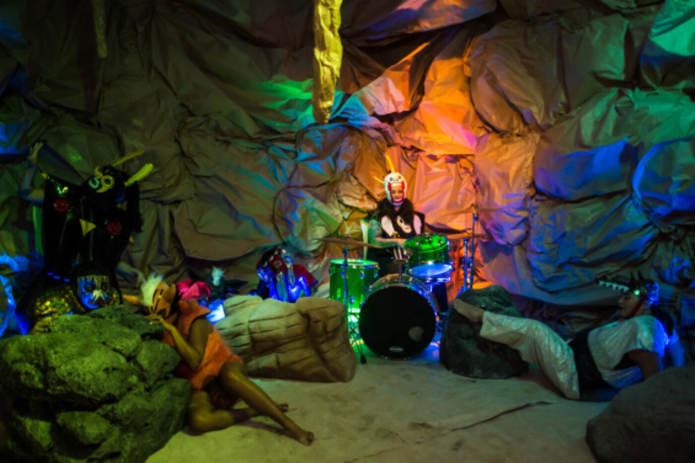 Photograph of a theatrically lit interior that looks like a cave. A performer is playing a drum kit, while a performer is draped over a large rock at left.